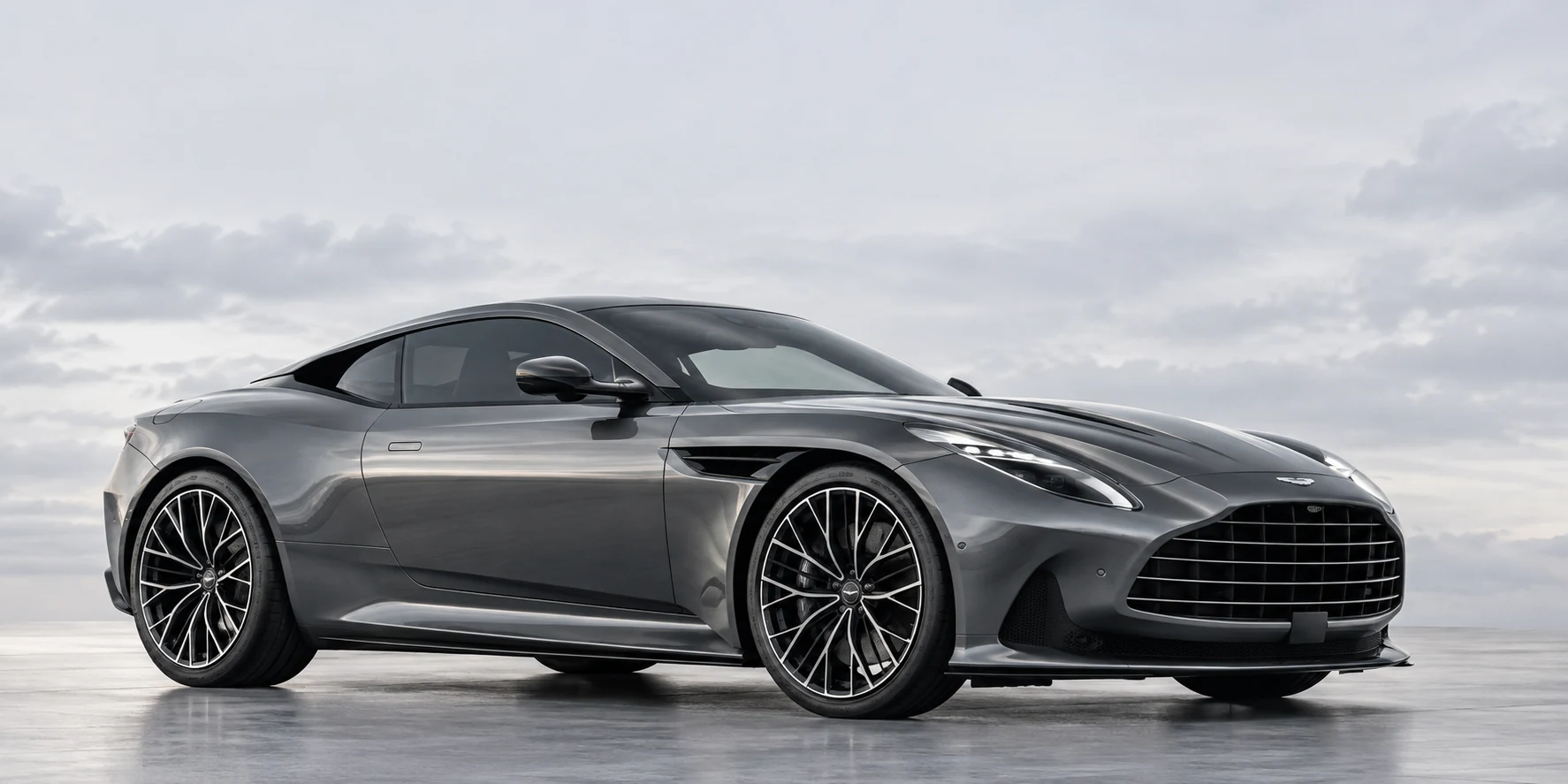 A graphite Aston Martin grand tourer photographed from a low angle beneath a pale, cloudy sky
